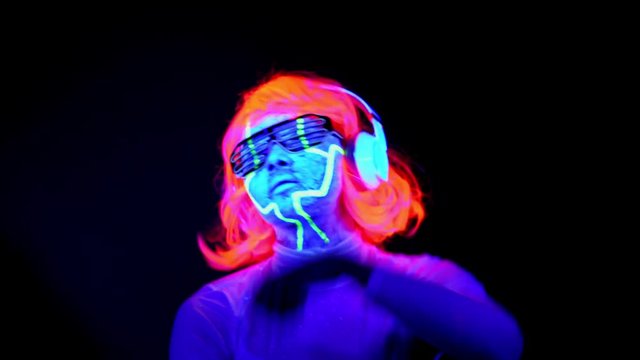 Beautiful sexy woman with UV cyborg face paint, wig, glowing glasses, clothing dancing and listening to music with headphones. Asian woman. Party concept.