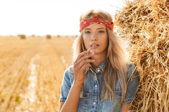 Image of gorgeous woman 20s standing near big haystack in golden field, and smoking cigarette during sunny day