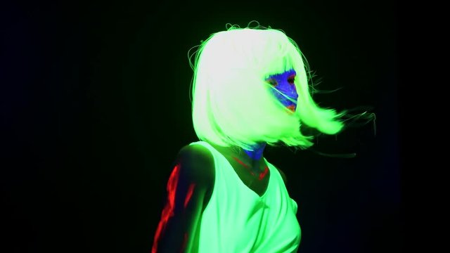 Beautiful sexy woman with UV face paint, wig, glowing clothing dancing in front of camera and shaking head, shoulder shot. Caucasian woman. Party concept.