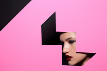 Face of young beautiful girl with a bright make-up and red lips looks through a hole in pink paper