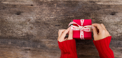 Christmas gift giving - someones hand in red knitted sweater making bow on red box with present,...