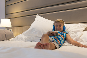 Happy small boy listening music over headphones while relaxing on bed.