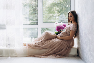 Sensetive portrait of pregnant woman. Expecting lady lies on the windowsill in a bright room