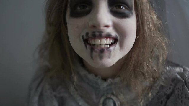 Girl in the costume of Halloween, in the makeup of the dead man screams and looks terribly at the camera. On the child's head is a wreath of skulls and a gray veil. Dynamic video, close-up