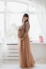 Beautiful pregnant woman in long beige dress standing in a bright cosy room with large windows