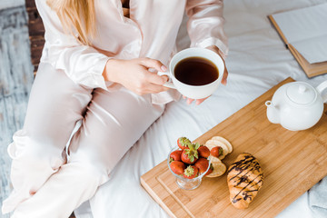 cropped image of woman in pajamas holding cup of tea in bed in morning