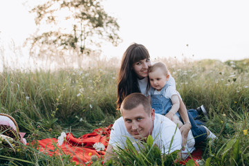 Fototapeta premium Family portrait. Mom, dad and their little daughter rest on a red plaid on a green summer field