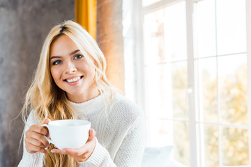 cheerful beautiful woman in sweater holding cup of coffee and looking at camera in bedroom in...