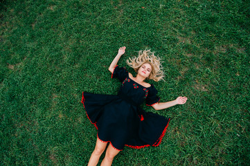 Odd strange unusual beautiful blonde woman in black ethnic linen dress lying on green grass....