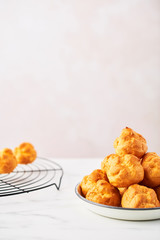 Bunch of fresh cream puff cakes on a white plate and wire rack on white marble table over pink background with copy space. Selective focus.