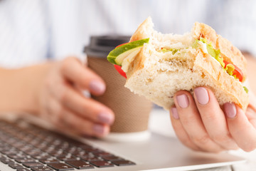 Woman eating a breakfast sandwich and drinking coffee while working with a laptop