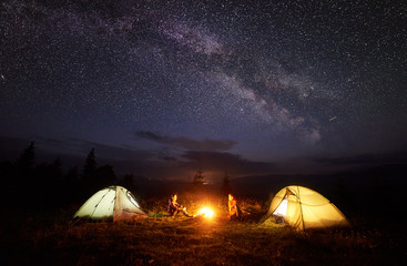 Fototapeta na wymiar Camping in mountains at night. Bright bonfire burning between two hikers, boy and girl sitting opposite each other near illuminated tents under beautiful evening starry sky and Milky way