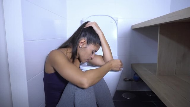 Attractive young and sad bulimic young woman feeling guilty and sick eating while sitting on the floor next to the toilet in eating disorders anorexia and bulimia concept.
