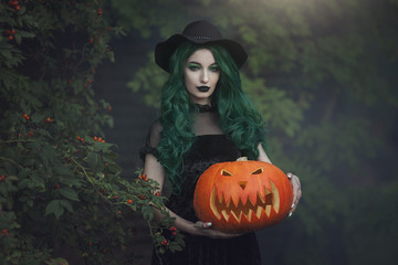 A Halloween witch with green hair is holding a carved scary pumpkin. Beautiful young woman in black...