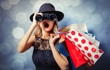 Portrait of a young style girl in black dress and hat with shopping bags and binoculars on gray background with bokeh