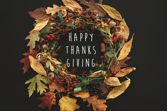 Happy Thanksgiving text on autumn wreath flat lay. Fall leaves circle with berries, nuts, acorns, flowers,herbs on black dark background. Seasons greetings card.