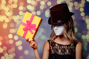 Portrait of a young style girl in top hat and white mask with gift box on gray background
