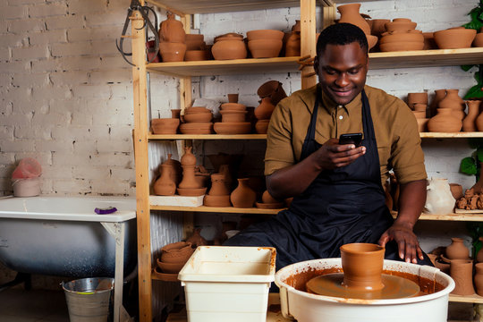 mixed race afro male potter with black apron and stylish dark shirt sitting at workshop table potter's wheel with handmade clay brown pot vase photographing himself portrait selfie on smartphone