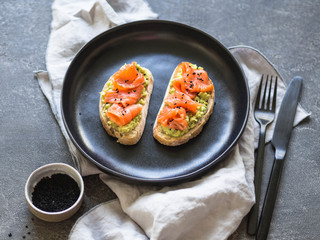 Toasts with avocado and salted salmon and black sesame seeds on a black plate on a gray background