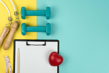 top view of the green dumbbells and jump rope, vinyl tape measure, a red heart symbol and black clipboard with pencil on the yellow and blue background