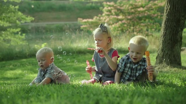Happy three kids blowing soap bubbles in park seatting on the grass Slow motion Ful HD