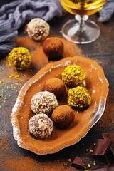 Variety of homemade dark chocolate truffles with cocoa powder, pistachios, almonds in dark brown background texture. Top view, copy space.