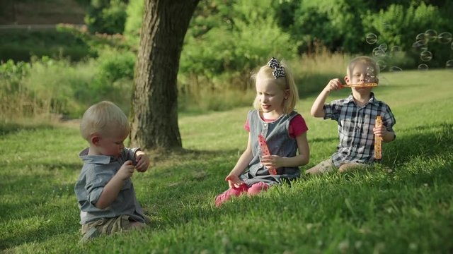 Happy three kids blowing soap bubbles in park seatting on the grass Slow motion Ful HD