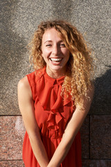 Young redgead curly hairstyle girl, weared in red dress, laughing, standing near granite wall