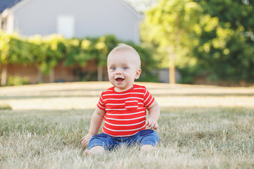 Cute adorable little Caucasian baby boy in red t-shirt and pants sitting in field meadow outside. Little happy smiling child in summer park on sunset. Lovely charming male infant having fun.