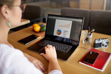 Close-up rear view of young business or student woman working at cafe with laptop computer, typing, looking at screen with statistic, using touchpad. View over the shoulder. Telecommuting concept.