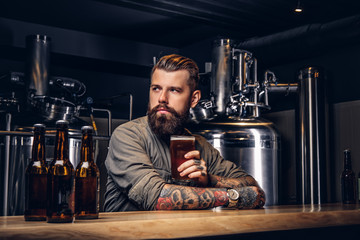 Tattooed hipster male with stylish beard and hair drinking beer sitting at the bar counter in the indie brewery.