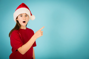 Fototapeta na wymiar Shocked Christmas Girl Wearing Santa Hat, Pointing to Copy Space