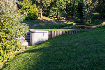 The bridge of the wooden pillars. Green lawn on the slope. A small pond in the background. Green trees in the park.