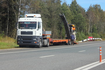 Low-frame european semi-trailer truck unloads asphalt remover summer day on the background of the roadside and green forest - transportation of special equipment for road repairs, transport logistics