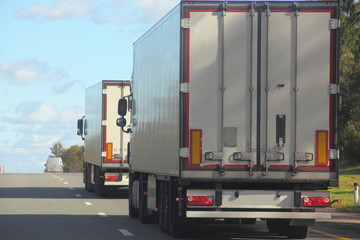 White trucks with a semi-trailers rides on a suburban highway road on a summer day against the blue sky and green forest, rear side view of the trailer - export transportation, transport logistics