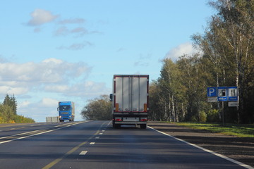 White truck with a semi-trailer rides on a suburban highway road on a summer day against the road signs, blue sky and green forest, rear view of the trailer - transportation, transport logistics, work