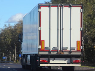 Heavy duty transport in logistics - white semi-trailer truck rides on asphalt highway in summer against green trees, road signs and blue sky, rear-side view