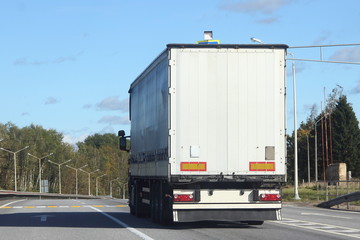 White truck with a semi-trailer go on a europe suburban highway road on a summer day against the blue sky and green forest, rear side view of the trailer - delivery, trade, transportation logistics