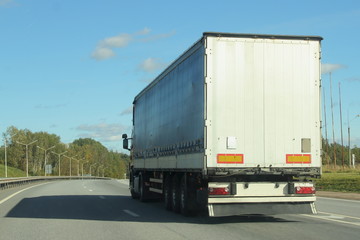 Beautiful white truck with a semi-trailer rides on turn of a suburban highway road in summer day against blue sky and green forest, side rear view of trailer - transportation, transport logistics