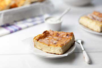Tender, airy casserole of cottage cheese, is a piece on a plate, next to the baking sheet, sour cream in a gravy boat, standing on a table, on a light background, side view