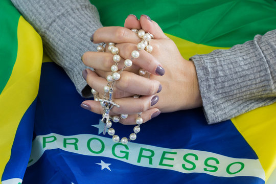 Close-up young female hand holding a rosary of pearls, praying over the Brazilian flag. Chaplet and faith. (Translation: "progress")