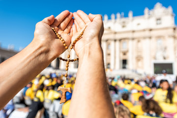 Macro closeup of hands holding wooden handmade Italian cross Catholic Assisi rosary with bokeh background of Vatican church St Peter's Square Basilica during mass on sunny day - obrazy, fototapety, plakaty