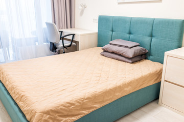 Closeup of minimalism bed with green blue turquoise headboard, office table desk chair, lamp, pillows in bedroom in staging model home, house or apartment by window balcony hotel