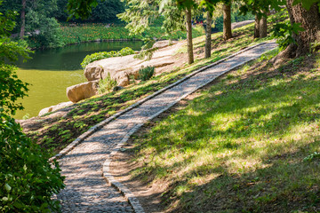 City park in the summer. Empty stone path by the lake in the shade of trees.