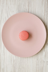 Pink macaroon on a pink plate over white wooden background, overhead view. Close-up. Flat lay, from above, top view.