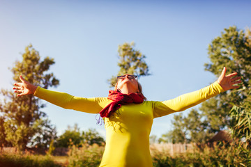 Middle-aged woman feeling free and happy on river bank on autumn day. Senior lady raising hands up