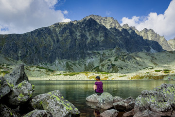 Redhead girl is sitting on the stone near beautiful mountain lake with huge rocks on the background
