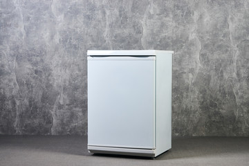 White Empty mini fridge or mini bar against gray textured wall background. International Exhibition furniture elements in large warehouse interior.