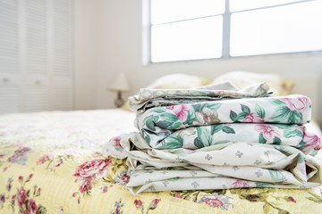 Stack of bedroom linen with floral, flower pattern on top of bed with quilt, quilted comforter,...