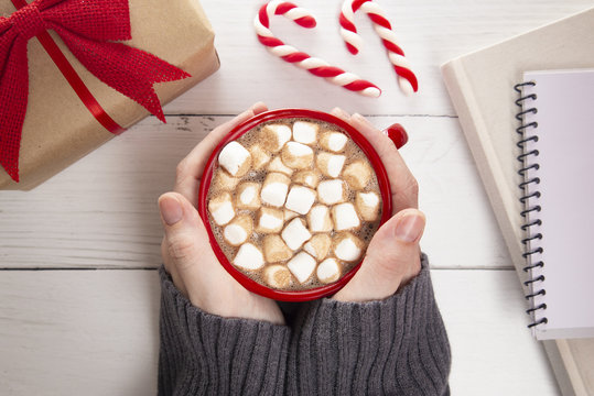 Person Holding a Hot Chocolate with Marshmallows on a Wooden Table with a Gift and a Novel to Read